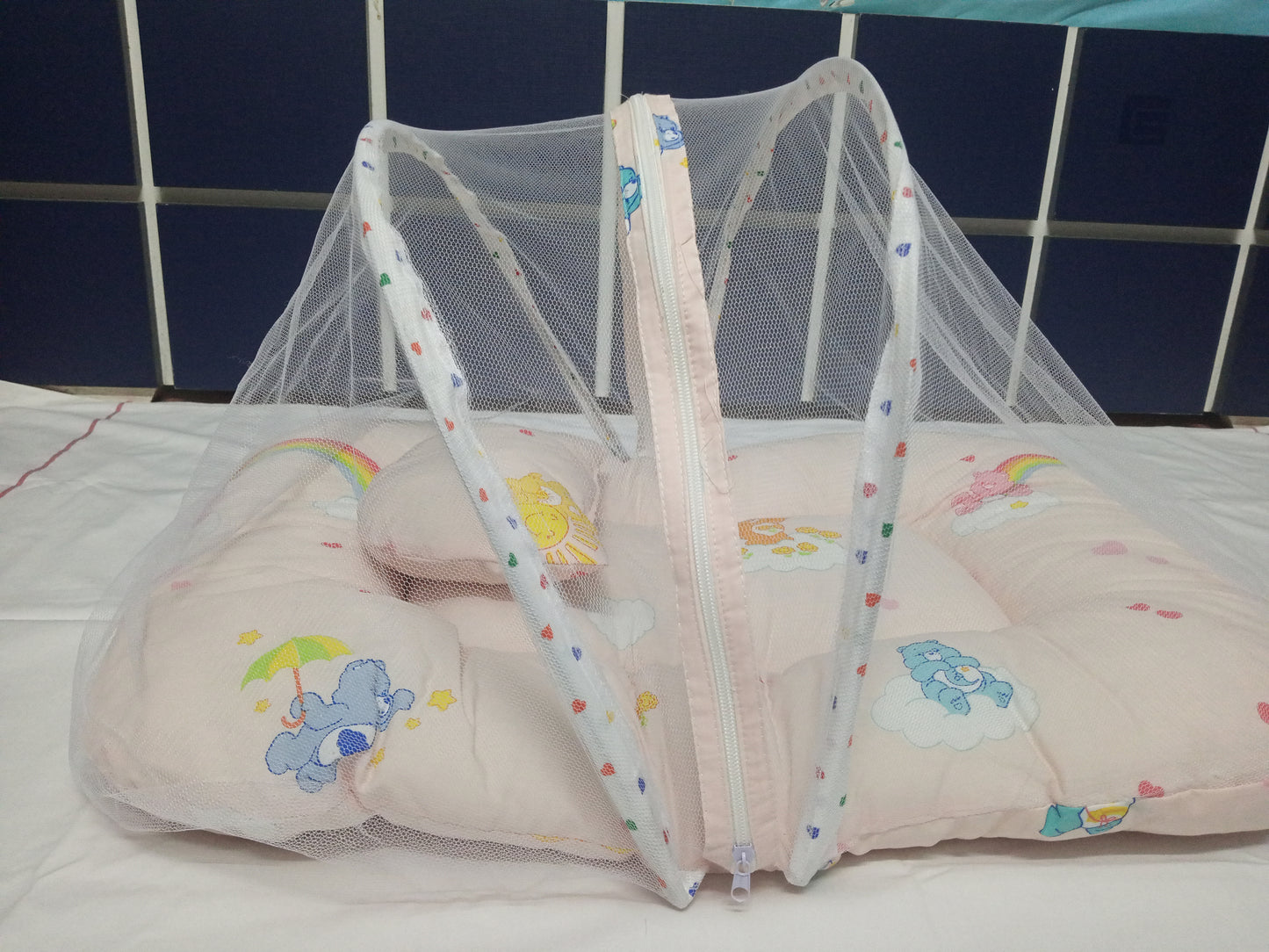 Baby crib with a pink mattress and colorful bear design against a blue tiled wall.