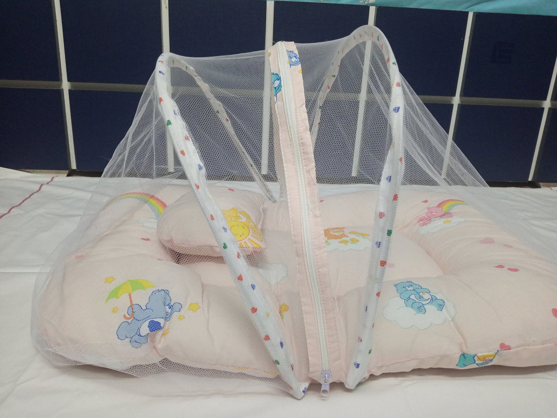 Baby crib with a pink mattress and colorful bear design against a blue tiled wall.